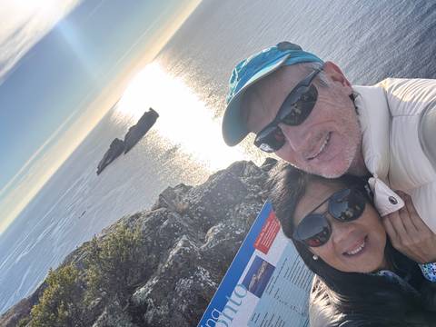       Couple takes a sunny selfie atop seaside cliffs with sparkling ocean and islands behind them.
  