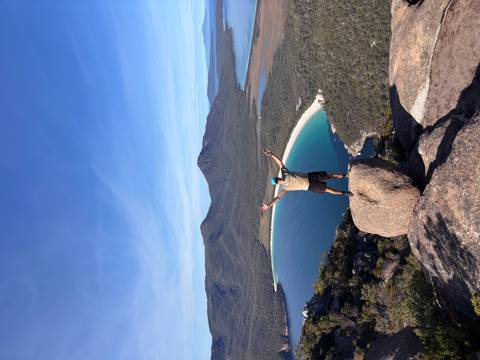       Adventurer with arms raised stands on a boulder high above the turquoise curve of Wineglass Bay.
  