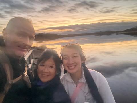       Blurred sunset selfie of three travellers beside calm water reflecting pastel clouds.
  