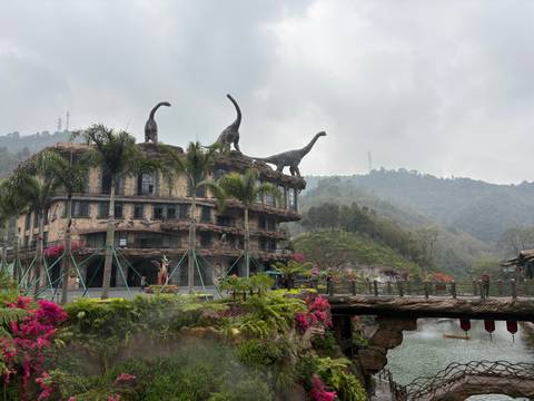       Themed building with dinosaur statues on roof, tropical plants, bridge and misty hills behind
  