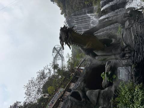       Large dinosaur statue in misty theme park with waterfall backdrop and observation deck
  