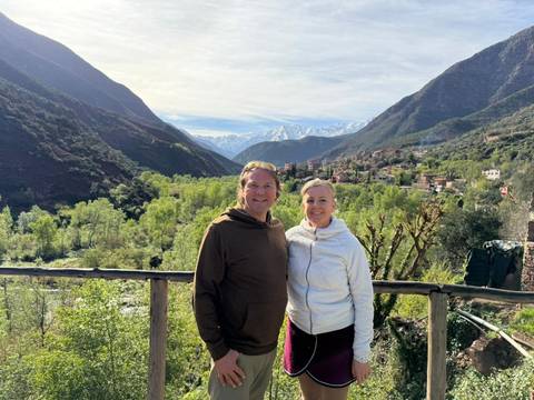       A smiling couple pose on a wooden railing with lush green valley and snowy Atlas peaks behind.
  