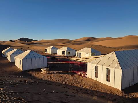       Rows of white canvas tents form a desert camp among rolling golden dunes under a cloudless sky.
  
