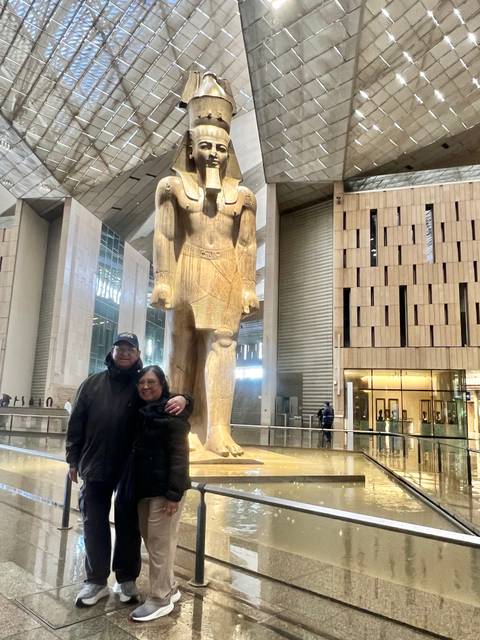       A couple stands beside a colossal pharaonic statue inside the Grand Egyptian Museum’s airy hall.
  