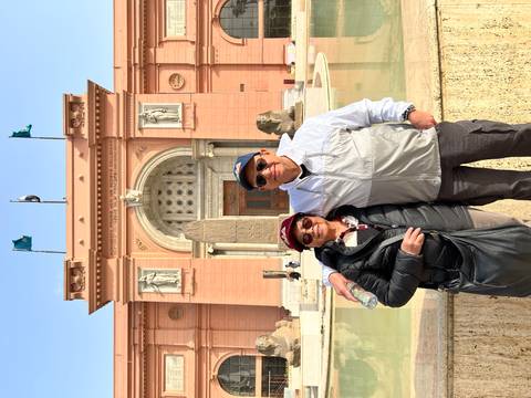       Visitors smile outside the pink-facade Egyptian Museum with ancient statues and a reflecting pool.
  