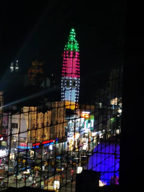       Night city scene of a brightly lit tower viewed through a mesh fence, blurry and noisy.
  