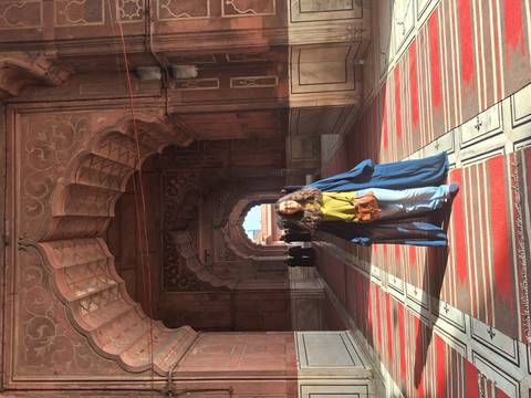       Traveler in long robe stands beneath ornate arches of a red sandstone mosque corridor
  