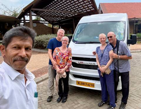       Driver takes a selfie with four smiling travellers standing beside a white tourist van in a parking area.
  