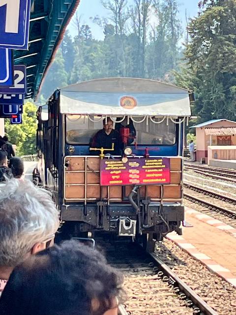       Blue heritage mountain railway engine arrives at a small hill station while passengers watch.
  