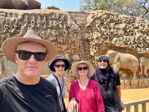       Four travellers pose in front of the vast Arjuna's Penance rock relief under bright sun.
  