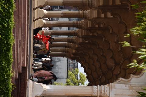       People move beneath a corridor of scalloped Mughal arches with a woman in a bright red dress twirling.
  