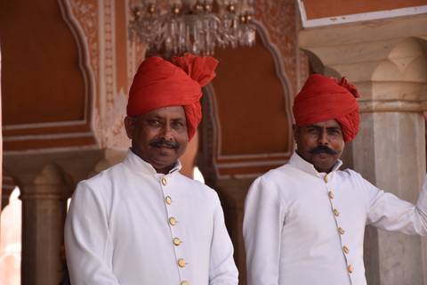       Two uniformed doormen wearing red turbans pose and smile inside an ornate palace corridor.
  