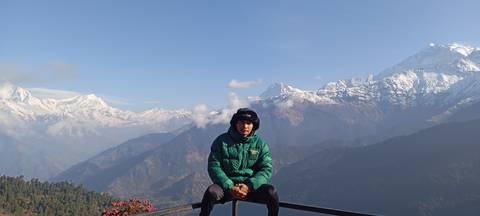      Young man in green down jacket sits on a railing with snow-capped Annapurna range behind.
  