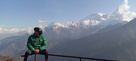       Traveller gazes to the side while seated on a balcony rail with panoramic snowy peaks.
  