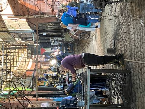       Metalworking artisans labor in narrow covered workshop aisle lit by lamps.
  