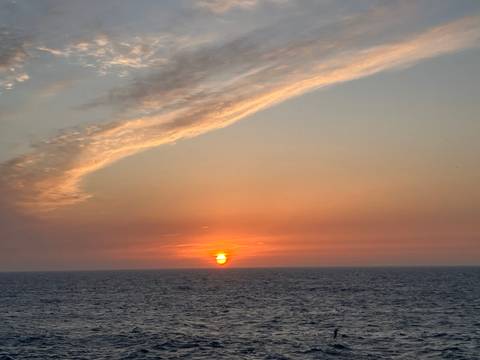       Brilliant orange sun sets over calm ocean horizon beneath sweeping clouds.
  