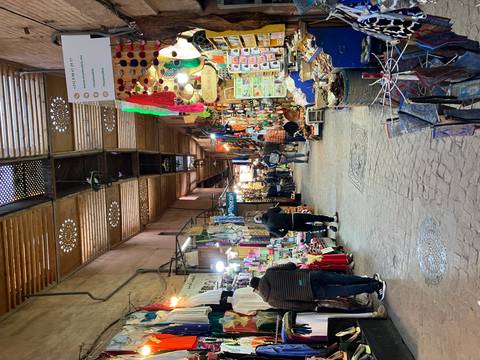       Covered market alley with colorful stalls, hanging lamps and a few shoppers in a Moroccan souk.
  