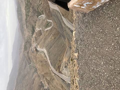       High-altitude serpentine road winding through barren brown mountains on an overcast day.
  