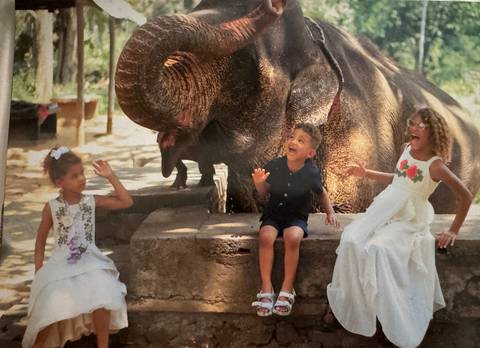       Three children dressed up and reacting joyfully beside a large elephant at an outdoor encounter.
  