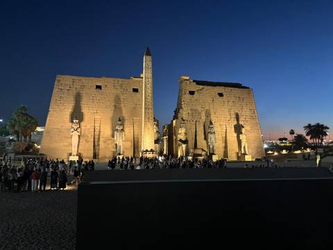       Evening view of illuminated Luxor Temple pylons and obelisk with crowds gathered below.
  