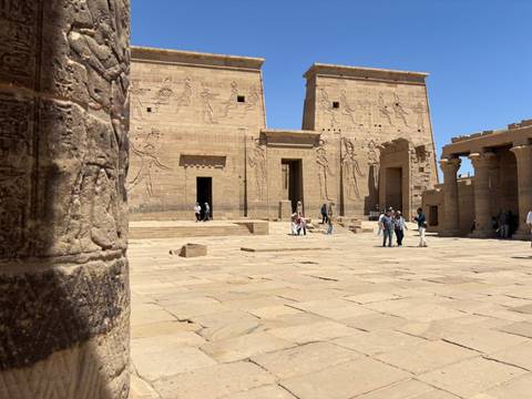       Open courtyard of Philae Temple with carved pylons and a few visitors exploring.
  