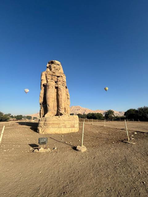       Colossi of Memnon statue standing tall with hot air balloons floating in the blue sky behind.
  