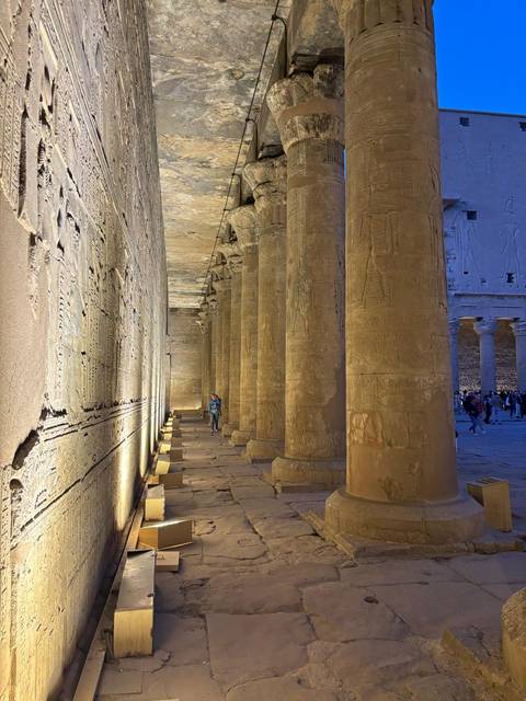       Rows of illuminated columns inside a Nile-side temple at dusk with visitors strolling.
  