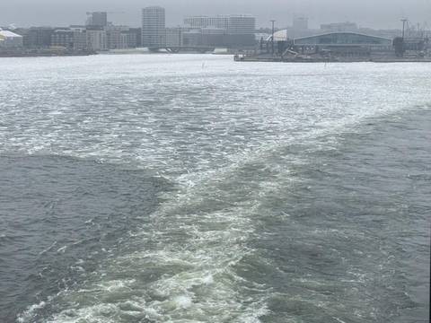       Choppy, partially frozen grey water with a foamy boat wake under an overcast sky and distant bridge.
  