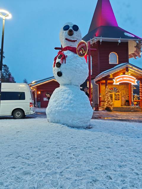       Large snowman outside a illuminated hotel reception with two smiling travellers peeking from behind.
  