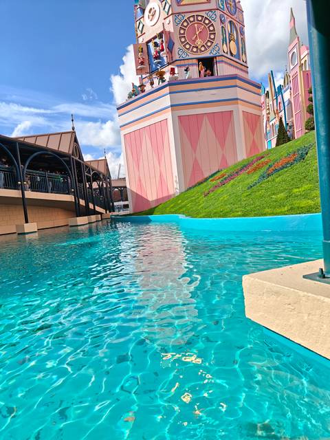       Bright turquoise canal in an amusement-park setting with arched bridge, pastel buildings and queuing visitors.
  