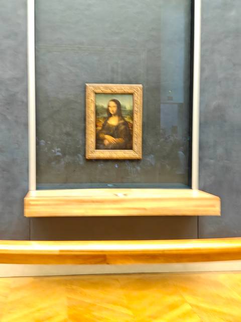       Framed Mona Lisa painting behind protective glass at a museum gallery with blurred crowd reflections.
  