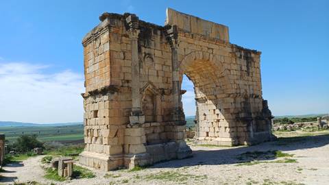       Massive triumphal stone arch standing amidst grassy plains at an expansive archaeological site.
  