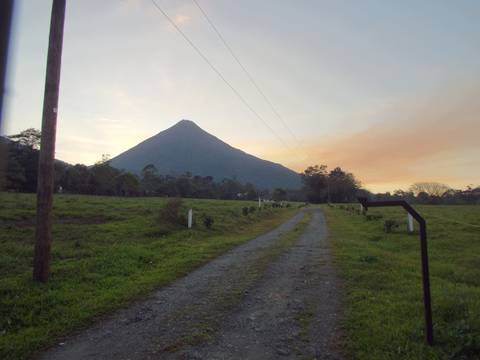      Conical volcano rising above grassy pasture at dusk with pastel sky hues and country road.
  