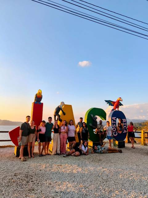       Cheerful tour group posing beside colourful 'Costa Rica' viewpoint sign adorned with parrot sculptures at sunset.
  