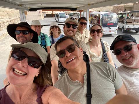       Smiling travellers taking a group selfie while seated on an open shuttle cart
  