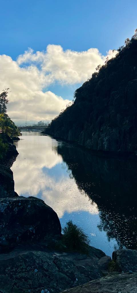       Calm river gorge reflecting cloudy sky with distant bridge and cliffs
  