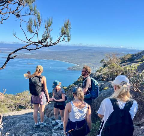       Small group of hikers resting on rocky lookout with sweeping view across turquoise bay, forested peninsula and distant mountains
  
