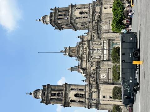       Front façade of Mexico City Metropolitan Cathedral with a police van parked in the plaza below a blue sky
  