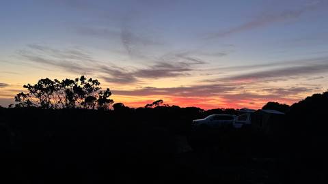       Silhouetted campsite with vehicle and trailer beneath a vivid pink and orange outback sunset sky
  
