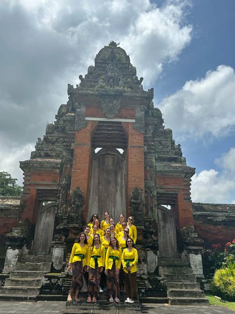       Travel group stands at a traditional red-brick Balinese temple gate under a cloudy sky.
  