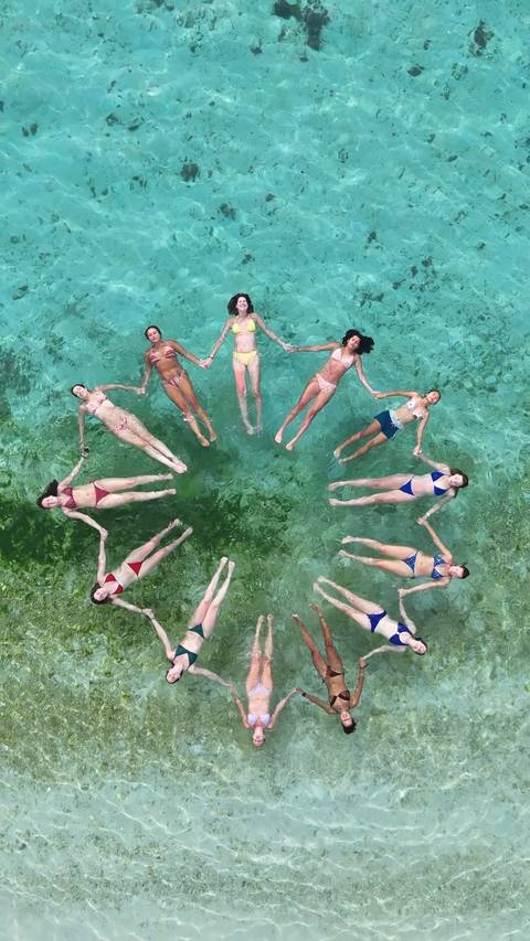       Aerial shot of women floating in a circle holding hands in clear turquoise sea.
  