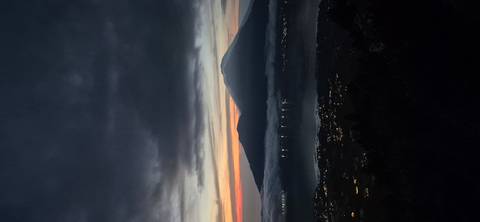       Silhouetted volcano rises above a lake and town lights at dawn with colorful clouds.
  