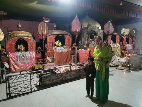       Woman in a green sari and a young boy pose in front of brightly decorated Hindu shrine statues inside a temple hall.
  