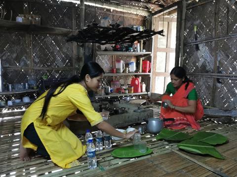      Two women in a rustic bamboo kitchen prepare food and set out banana-leaf plates.
  
