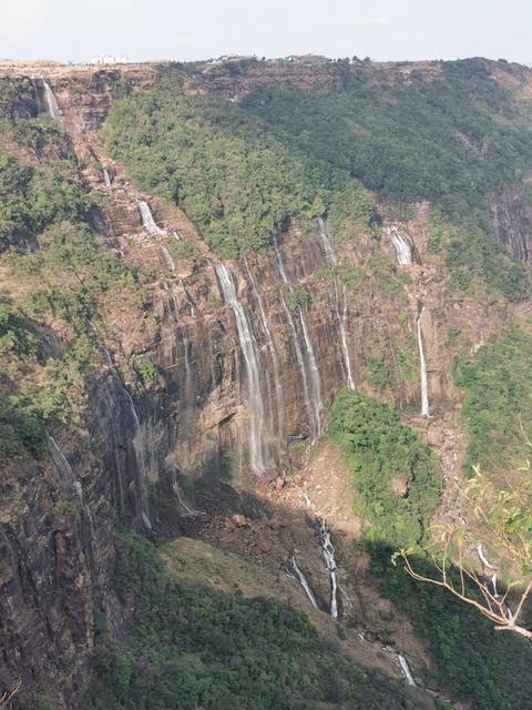       Series of tall, narrow waterfalls cascade down a steep rocky cliff covered with green vegetation.
  