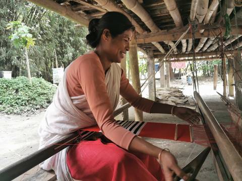       Smiling woman works a traditional handloom beneath a bamboo shelter in a rural setting.
  