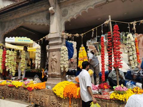       Busy flower market stall draped with colorful garlands and marigolds while vendors arrange blooms.
  