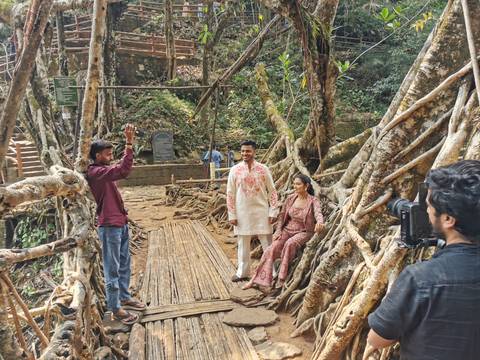       Couple poses for photos on a living root bridge walkway while friends capture the moment in a forested gorge.
  