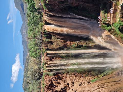       Powerful twin waterfalls plunging over a tall cliff into a lush canyon framed by mountains and blue sky
  