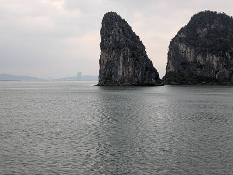       Overcast view of a tall limestone karst rising from calm gray waters with distant city skyline in Halong Bay.
  